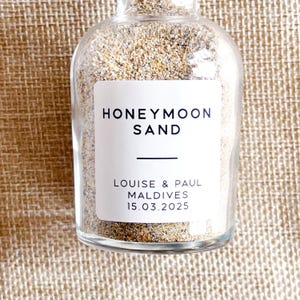 May include: A clear glass bottle filled with sand, with a white label reading "HONEYMOON SAND." The label also states "LOUISE & PAUL MALDIVES 15.03.2025." The sand inside is a mix of beige and brown.