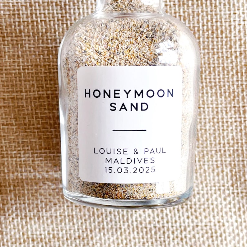 May include: A clear glass bottle filled with sand, with a white label reading "HONEYMOON SAND." The label also states "LOUISE & PAUL MALDIVES 15.03.2025." The sand inside is a mix of beige and brown.