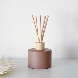 May include: A brown ceramic essential oil diffuser with wooden reeds. The diffuser has a cylindrical base and a light wood collar holding the reeds. The reeds are light brown and arranged in a fan shape.
