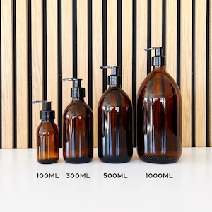 May include: Four amber glass bottles with black pump dispensers. The bottles are labeled with their volume: 100ML, 300ML, 500ML, and 1000ML. The bottles are arranged in a row on a white surface, with a wooden slatted background.