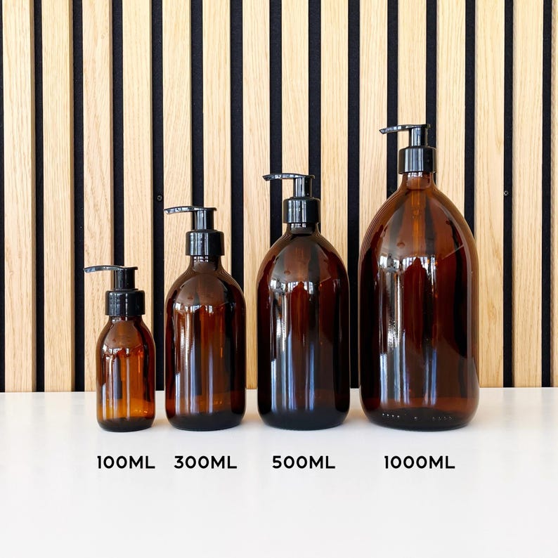 May include: Four amber glass bottles with black pump dispensers. The bottles are labeled with their volume: 100ML, 300ML, 500ML, and 1000ML. The bottles are arranged in a row on a white surface, with a wooden slatted background.