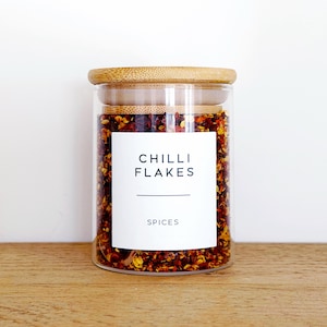 May include: A glass jar filled with red chilli flakes, with a bamboo lid. The jar has a white label that reads "CHILLI FLAKES" and "SPICES". The jar is on a wooden surface.