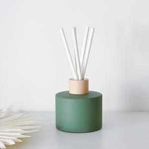 May include: A cylindrical matte forest green glass reed diffuser bottle with a light-coloured wooden collar at the neck. Five slender white reed sticks are inserted into the opening, extending upwards in a fan-like arrangement from the centre.