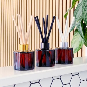 May include: Three amber glass reed diffusers with gold, black, and silver collars. Each diffuser holds a different colour of reeds: natural, black, and white. The diffusers sit on a white shelf with a wooden slatted background.