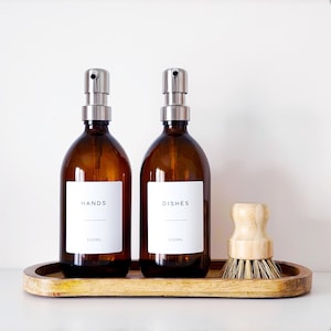 May include: Two brown glass bottles with silver pump dispensers, labelled "HANDS" and "DISHES", on a wooden tray, with a wooden dish brush. Each bottle holds 500ml. Ideal for a kitchen or bathroom.