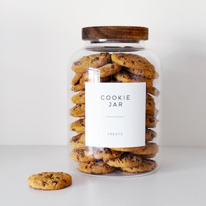 May include: A glass cookie jar filled with chocolate chip cookies. The jar has a wooden lid and a white label that reads "COOKIE JAR TREATS". One cookie sits in front of the jar.