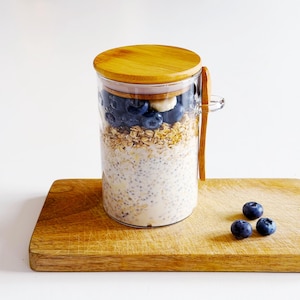 May include: A clear glass jar with a wooden lid filled with layers of oats, chia seeds, blueberries, and a banana slice. A wooden spoon rests on the side. The jar sits on a wooden chopping board, with three blueberries nearby.