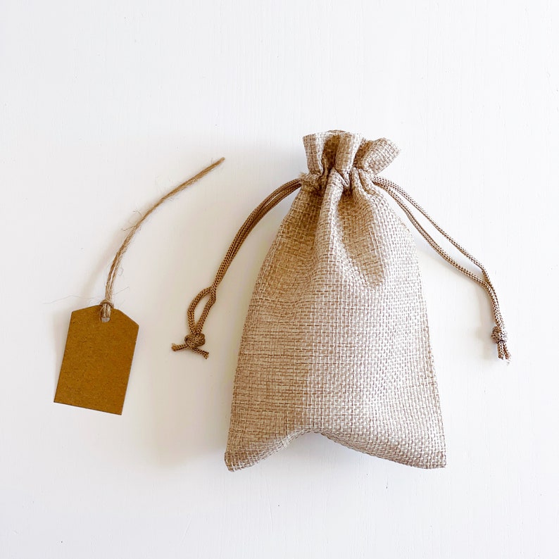 May include: A small, beige burlap drawstring bag with a matching blank tag attached by twine. The bag is closed at the top with a drawstring. The tag is rectangular with rounded corners.