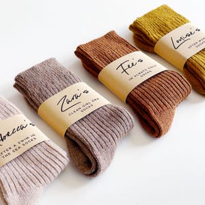 May include: Four pairs of ribbed knit socks in light beige, mauve, rust orange, and mustard yellow. Each pair is wrapped in a tan paper band with black text. The labels read Rebecca's, Zara's Clean Girl Era, Fee's I'm Always Cold, and Louise's After Work.