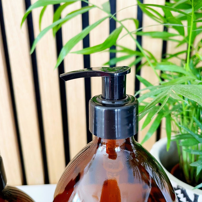 May include: Amber glass bottle with a black pump dispenser. The bottle has a rounded shape and a glossy finish. The pump is made of black plastic. The background includes a wooden slatted wall and green plants.