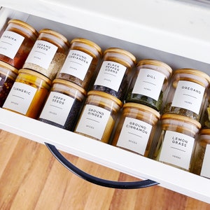 An open kitchen draw with lots of small jars lined up, laying down neatly. Each glass jar has a wooden bamboo lid and an airtight seal. On the front of each jar is a white label printed in a minimalist design with the name of a herb or spice on each.