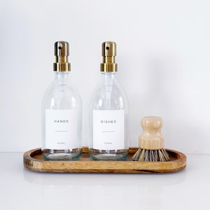 May include: Two clear glass soap dispensers with gold-toned metal pumps, each featuring a white label printed with the words HANDS or DISHES and 500ML. A wooden dish brush with natural bristles sits beside them on an oval wooden tray.