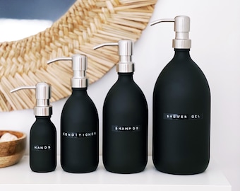 May include: Four black matte soap dispensers with silver pump tops. The dispensers are labelled "HANDS", "CONDITIONER", "SHAMPOO", and "SHOWER GEL". They are arranged on a white shelf, with a woven mirror in the background.
