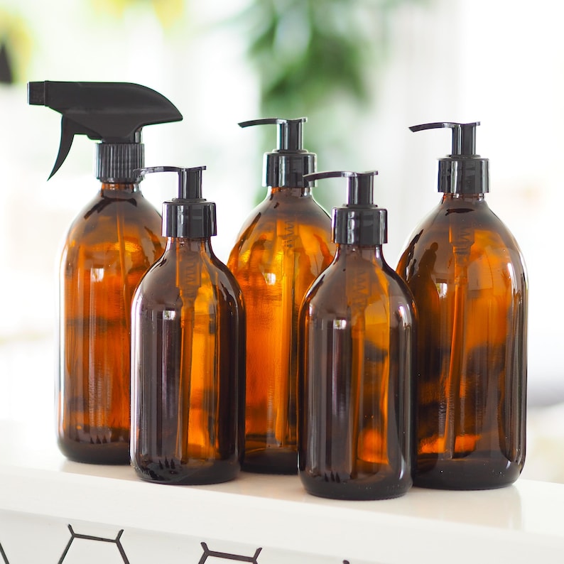 May include: A collection of amber glass bottles with black pump dispensers and a spray bottle. The bottles vary in size and shape, suitable for soaps, lotions, or cleaning solutions. The bottles are arranged on a white surface.