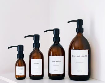 Puede incluir: Cuatro botellas de vidrio ámbar con dispensadores de bomba negros están dispuestas en un estante blanco. Las botellas están etiquetadas como "HANDS", "DISHES", "SHAMPOO" y "CONDITIONER". La botella de "CONDITIONER" es la más grande, con 1000 ml. Un abanico decorativo está en el estante.