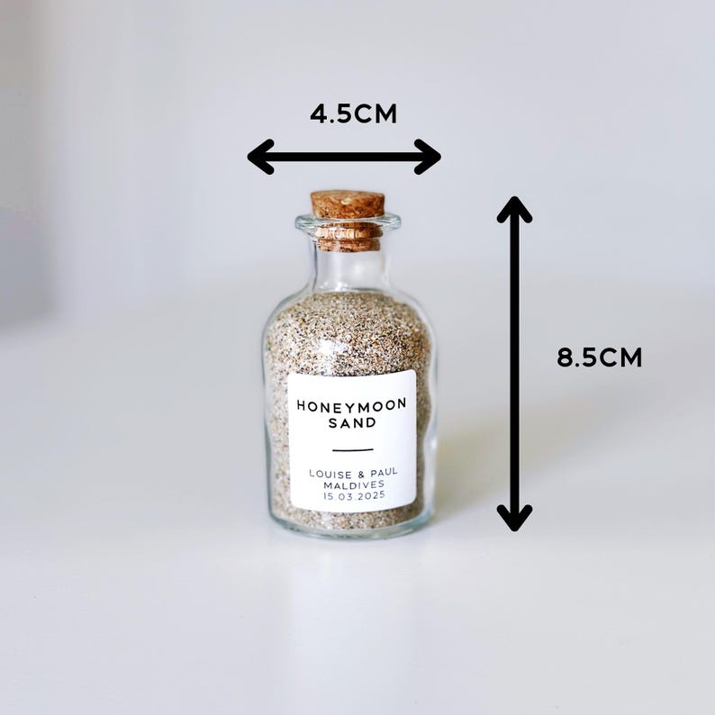 May include: A clear glass bottle filled with sand, measuring 8.5cm tall and 4.5cm wide, with a cork stopper. The label reads "Honeymoon Sand" with names and a date. A wedding keepsake.