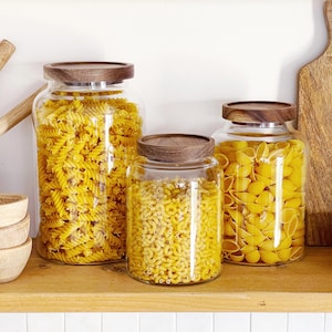 May include: Three glass jars with wooden lids, each containing a different pasta shape. The jars sit on a wooden shelf. The pasta varieties are fusilli, ditalini, and conchiglie, all in shades of yellow.