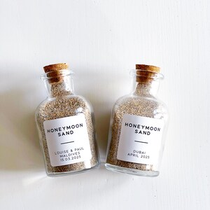May include: Two glass bottles filled with sand, each sealed with a cork stopper. White labels read "HONEYMOON SAND" with names and dates below. One reads "Louise & Paul Maldives 15.03.2025" and the other "Dubai April 2025".