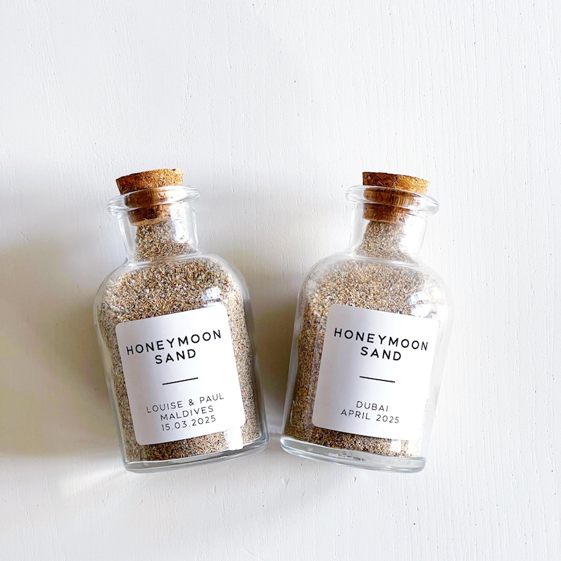 May include: Two glass bottles filled with sand, each sealed with a cork stopper. White labels read "HONEYMOON SAND" with names and dates below. One reads "Louise & Paul Maldives 15.03.2025" and the other "Dubai April 2025".