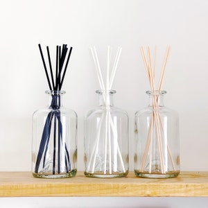 May include: Three clear glass bottles with different coloured diffuser sticks. The bottles are on a wooden shelf. The sticks are black, white, and beige.