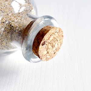 May include: A clear glass bottle filled with sand, sealed with a cork stopper. The sand is a mix of beige and tan colours. The cork is a light brown colour and has a textured surface.