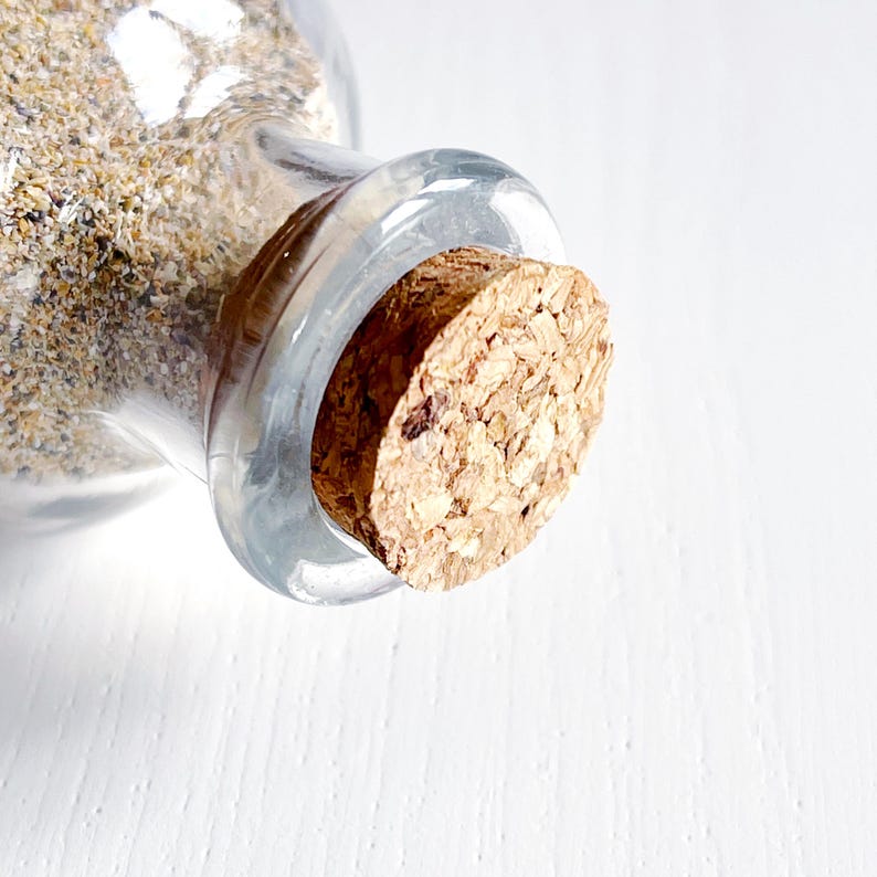 May include: A clear glass bottle filled with sand, sealed with a cork stopper. The sand is a mix of beige and tan colours. The cork is a light brown colour and has a textured surface.
