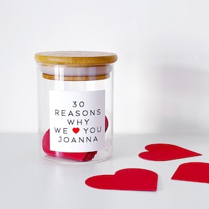 May include: A glass jar with a bamboo lid, filled with red heart-shaped notes. The jar has a white label with the text "30 REASONS WHY WE ❤️ YOU JOANNA". Three red paper hearts are placed nearby. The jar is on a white surface.