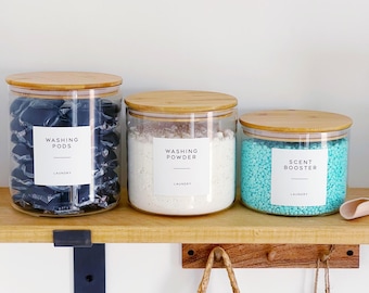May include: Three clear glass jars with bamboo lids on a wooden shelf. The jars contain laundry products: washing pods, washing powder, and scent booster. Each jar has a white label with black text. A woven basket with clothespins hangs below the shelf.