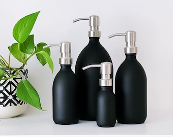 May include: Four black soap dispensers with silver pump heads are displayed. The dispensers are of varying sizes, with a small one at the front and a large one at the back. A plant with green leaves is on the left, in a patterned pot.