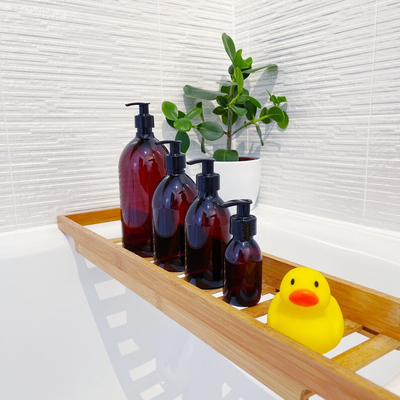 May include: A wooden bath tray holds four amber-colored pump bottles with black pumps, a small potted plant, and a yellow rubber duck. The bottles vary in size, suggesting a set for bath products.
