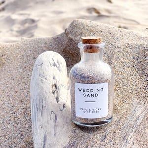 May include: A glass bottle filled with sand, sealed with a cork stopper. A white label reads "WEDDING SAND" with the names "PAUL & VICKY" and the date "10.05.2024". The bottle is on a piece of driftwood, with sand in the background.