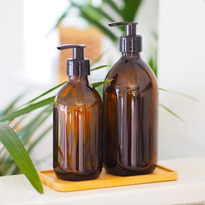 May include: Two amber glass soap or lotion dispensers with black pumps sit on a small bamboo tray. The bottles have a ribbed design and are different sizes. Green plant leaves are visible in the background.