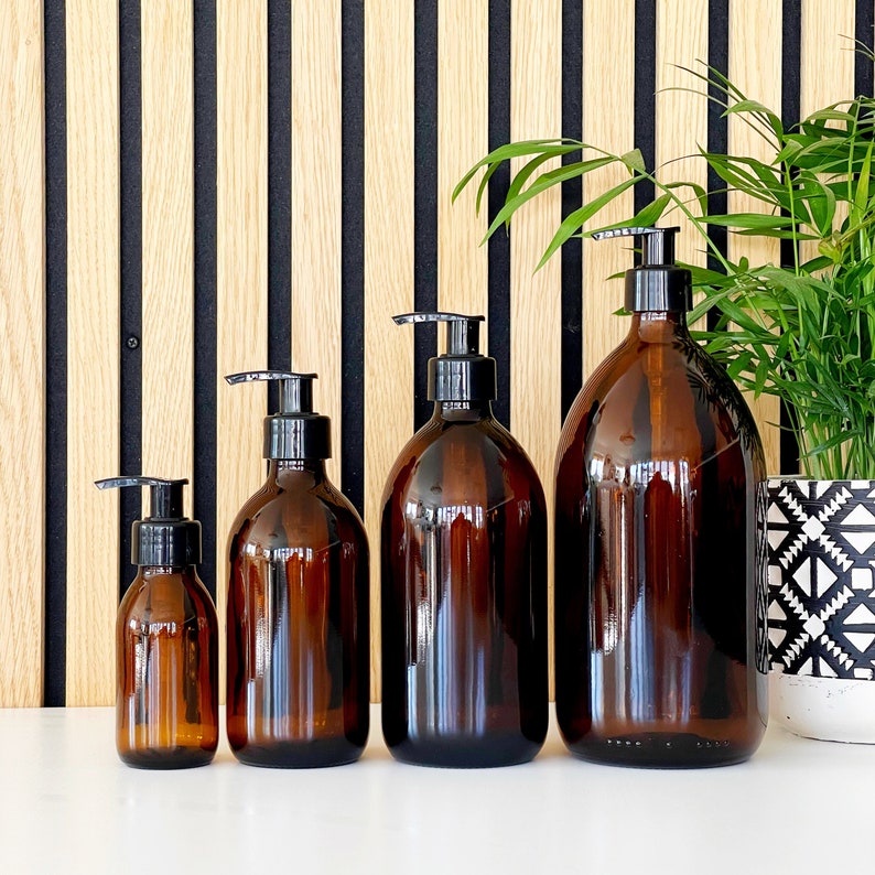 May include: Four amber glass bottles with black pump dispensers of varying sizes are arranged in a row. The bottles are on a white surface, with a wooden slat wall in the background. A potted plant is to the right.