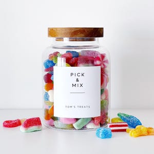 May include: A large glass jar with a wooden lid, filled with a variety of colourful sweets. The jar has a white label that reads "PICK & MIX" and "TOM'S TREATS". Several sweets are scattered around the jar, including red, blue, and yellow gummy sweets.