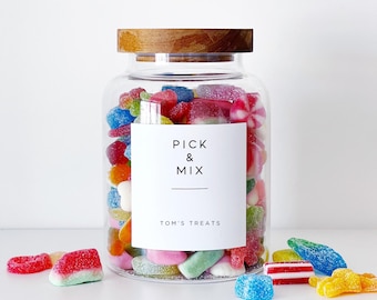 May include: A large glass jar with a wooden lid, filled with a variety of colourful sweets. The jar has a white label that reads "PICK & MIX" and "TOM'S TREATS". Several sweets are scattered around the jar, including red, blue, and yellow gummy sweets.