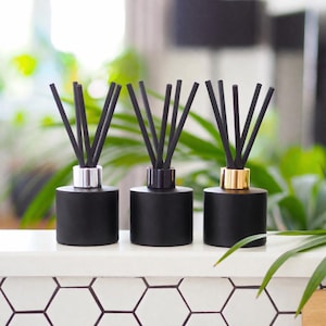 May include: Three black reed diffusers with silver, black, and gold collars. Each diffuser has several black reeds extending upwards. The diffusers are cylindrical and sit on a white surface with a hexagonal tile pattern.
