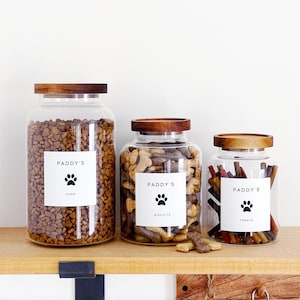 May include: Three glass jars with wooden lids, labelled "Paddy's" and paw prints, filled with dog food, biscuits, and treats. The jars are arranged on a wooden shelf, providing a neat storage solution for pet supplies.