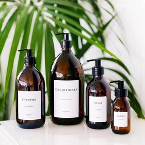 May include: Four amber glass pump bottles with black pumps and white labels. The labels read "Shampoo", "Conditioner", "Dishes", and "Hands". The bottles are arranged on a white surface, with a green leafy background.