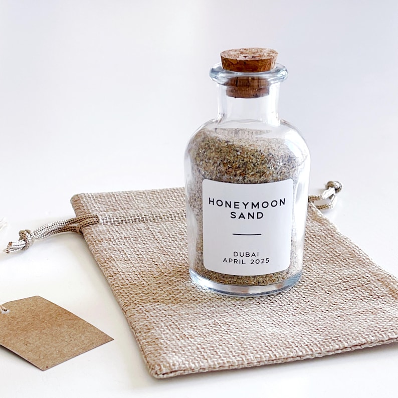 May include: A clear glass bottle filled with sand, labelled "HONEYMOON SAND DUBAI APRIL 2025", sits on a small hessian bag. The bottle has a cork stopper. A small, blank tag hangs nearby. The background is white.