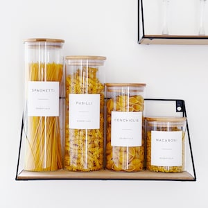 Four glass food storage jars on a shelf, on a wall.
The jars have wooden bamboo lids with airtight seals. The are 4 different sizes, each one has a different pasta in. On the front of each is a white waterproof minimalist personalised printed label.