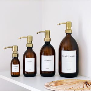 May include: Four amber glass bottles with brass-coloured pump dispensers. The bottles are labelled "HANDS", "DISHES", "SHAMPOO", and "CONDITIONER". The "SHAMPOO" bottle is 500ml, and the "CONDITIONER" bottle is 1000ml. The bottles are arranged on a white shelf.