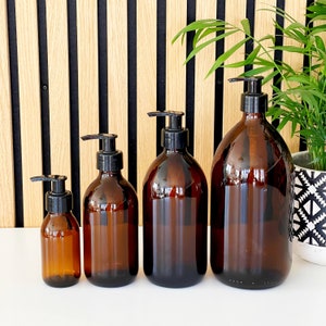 May include: Four amber glass bottles with black pump dispensers. The bottles vary in size, from small to large. The background features a wooden slat wall and a potted plant, creating a clean, modern aesthetic.