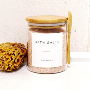 May include: A glass jar filled with pink bath salts, labelled "BATH SALTS BATHROOM." It has a wooden lid and a small wooden spoon. A natural sponge is to the left. The items are on a white surface.