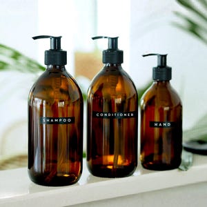 May include: Three amber glass bottles with black pump dispensers. The bottles are labelled "SHAMPOO", "CONDITIONER", and "HAND" in white text on black labels. The bottles are arranged on a white surface.