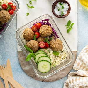 May include: A rectangular glass container with meatballs, cherry tomatoes, sliced cucumber, and cauliflower rice. Fresh herbs garnish the meal. The container rests on a marble board, with wooden cutlery visible.