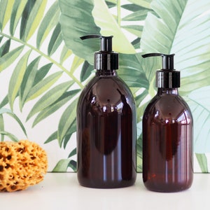 May include: Two brown, rounded bottles with black pump dispensers. The bottles are different sizes and sit on a white surface. A natural sponge and a tropical leaf pattern background are also visible.