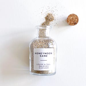 May include: A clear glass bottle filled with sand, labelled "HONEYMOON SAND." The label also includes the names "LOUISE & PAUL" and the date "15.03.2025." A cork stopper sits beside the bottle. Sand spills from the bottle's opening.
