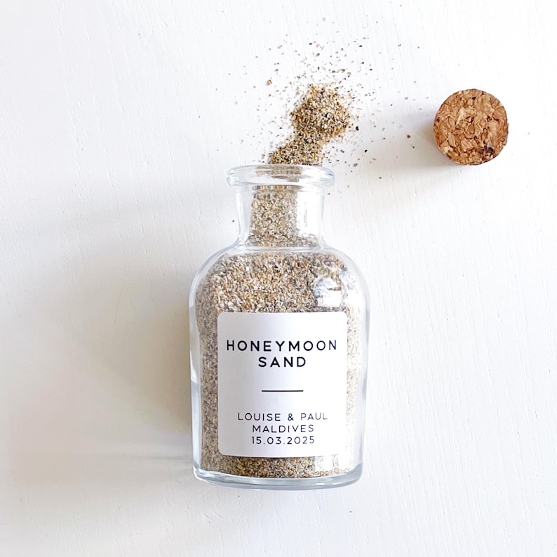 May include: A clear glass bottle filled with sand, labelled "HONEYMOON SAND." The label also includes the names "LOUISE & PAUL" and the date "15.03.2025." A cork stopper sits beside the bottle. Sand spills from the bottle's opening.