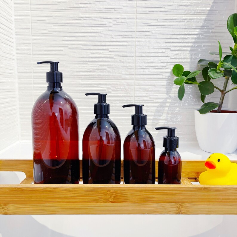 May include: Four amber-colored plastic pump bottles of varying sizes, arranged in a row. Each bottle has a black pump dispenser. The bottles are on a bamboo tray, with a rubber duck and a potted plant in the background.