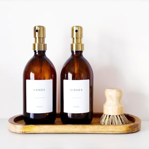 May include: Two amber glass bottles with brass pumps, labelled "HANDS" and "DISHES", on a wooden tray. A small wooden dish brush sits beside the bottles. Each bottle holds 500ml.